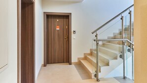 A hallway with a wooden door, a staircase with glass railings, and a light fixture on the ceiling.