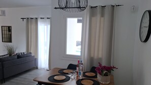 Ein modernes Wohnzimmer mit einer Couch, einem Esstisch mit Untersetzern, Stühlen, einer Topfpflanze und einer Wanduhr.