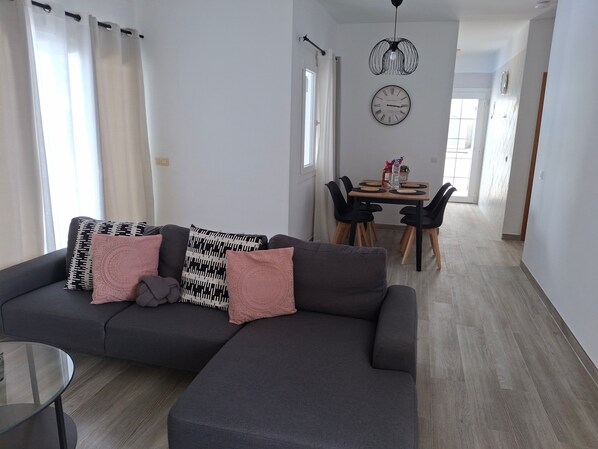 Een moderne woonkamer met een donkergrijze hoekbank, een glazen salontafel en een eetgedeelte met een houten tafel en zwarte stoelen.