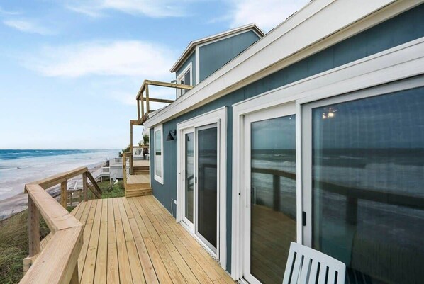 A coastal house with a wooden deck, large sliding glass doors, and a view of the ocean.