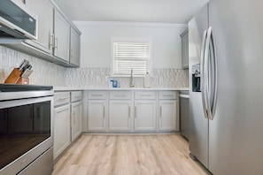 A kitchen with white cabinets, a stainless steel refrigerator, and a window with blinds.