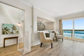 A spacious room with a large window overlooking the ocean, a dining area with a white table and chairs, and a staircase leading to an upper level.