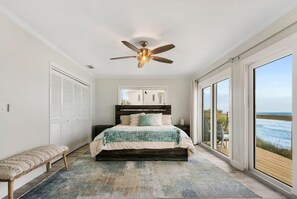 A bedroom with a large bed, a ceiling fan, a bench, and a view of the ocean through large windows.