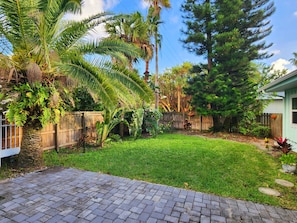 A backyard with a brick patio, a wooden fence, and various plants including palm trees and a large conifer. - Beach House with Ocean View, Heated Pool, and Rooftop Deck! Dog-friendly! (Cocoa Beach)