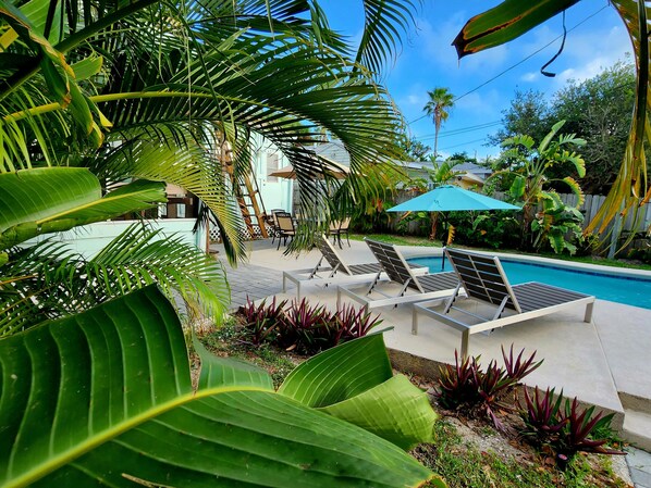 A poolside area with lounge chairs, a blue umbrella, and tropical plants. - Beach House with Ocean View, Heated Pool, and Rooftop Deck! Dog-friendly! (Cocoa Beach)