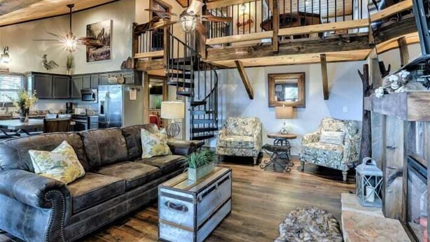 A spacious living area with a leather sofa, two patterned armchairs, a coffee table, and a rustic fireplace. A wooden staircase connects the living area to an upper level. - Smoky Mountain Loft - UNBELIEVABLE MOUNTAIN VIEWS, Great Location, Free Tickets! (Pigeon Forge)