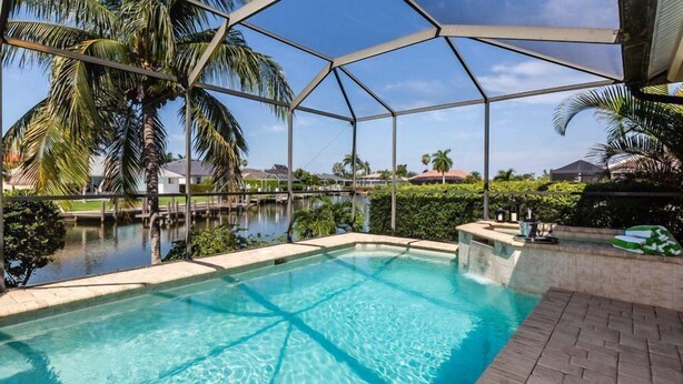 A swimming pool with a clear blue water, surrounded by a covered area with palm trees and a view of houses in the background. - Marco's Charmer: luxury coastal living, magical sunsets, tranquil water views, relaxing hot tub. (Marco Island)