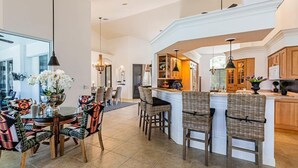 A modern kitchen with a central island, dining area with patterned chairs, and a large window. - Marco's Charmer: luxury coastal living, magical sunsets, tranquil water views, relaxing hot tub. (Marco Island)
