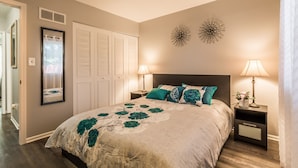 A bedroom with a bed, two bedside lamps, a nightstand, a mirror, a door with louvered shutters, and two wall decorations. - Comfortable and elegant 3BDR duplex, unit # 3 (Overland Park)
