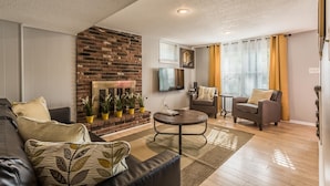 A living room with a brick fireplace, a flat-screen TV mounted above it, two armchairs, a coffee table, and a sofa. - Comfortable and elegant 3BDR duplex, unit # 3 (Overland Park)