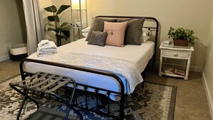 Ein ordentlich eingerichtetes Schlafzimmer mit Bett, Nachttisch, Stuhl und einer kleinen Ablage mit einer Pflanze.
