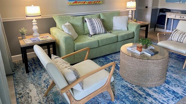 Ein Wohnzimmer mit einem grünen Sofa, zwei Sesseln, einem Couchtisch und einem Bild mit einer Strandansicht.