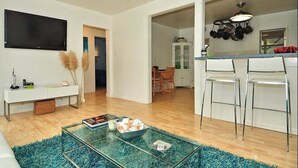 A modern living room with a glass coffee table, a flat-screen TV, and a dining area with a kitchen visible in the background.