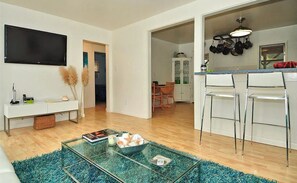 Uma sala de estar moderna com uma mesa de centro de vidro, uma televisão de tela plana e um espaço de jantar, com cozinha visível ao fundo.