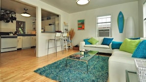 A modern living room with a white sofa, teal and green cushions, a coffee table, and a kitchen area with white cabinets and a bar stool.