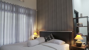 A neatly made bed with white linens and a black and white pillow, a bedside lamp, and a small table with a lamp and a tissue box. - Sayana Homestay (Sleman)
