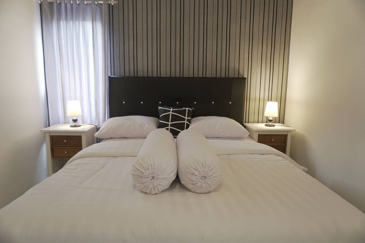 A neatly made bed with white linens and decorative pillows, two bedside tables with lamps, and a headboard with vertical lines.