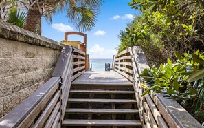A wooden staircase leading to a beach, surrounded by greenery and a clear sky.