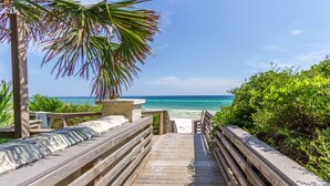 Een houten looppad dat naar een strand leidt, met een palmboom en begroeiing aan de zijkanten.