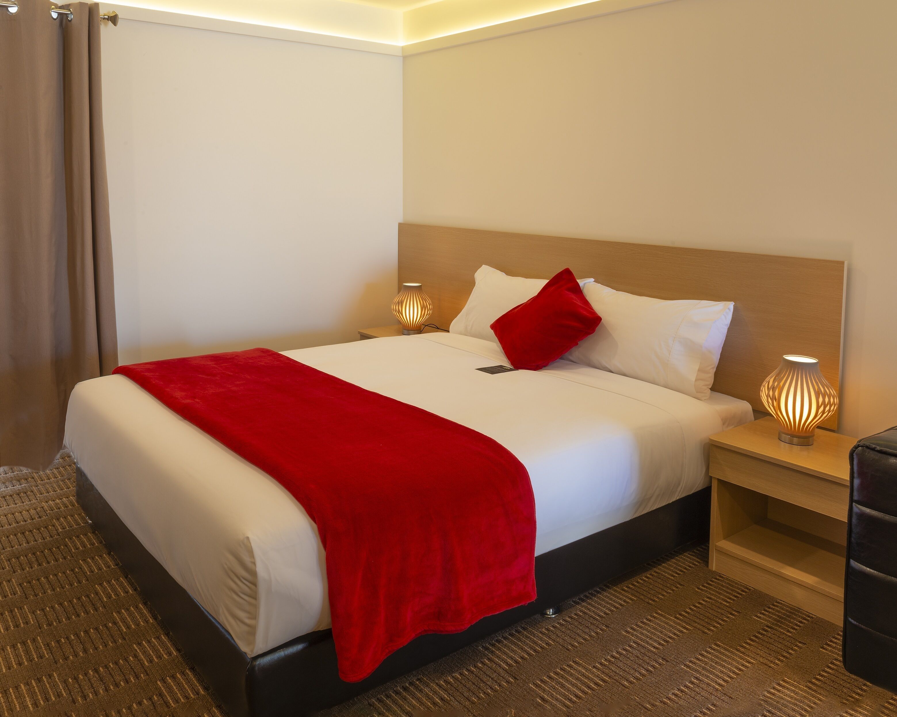 Een net opgemaakt bed met een rode plaid, witte kussens en een houten hoofdbord. Aan weerszijden van het bed staan twee nachtlampjes.