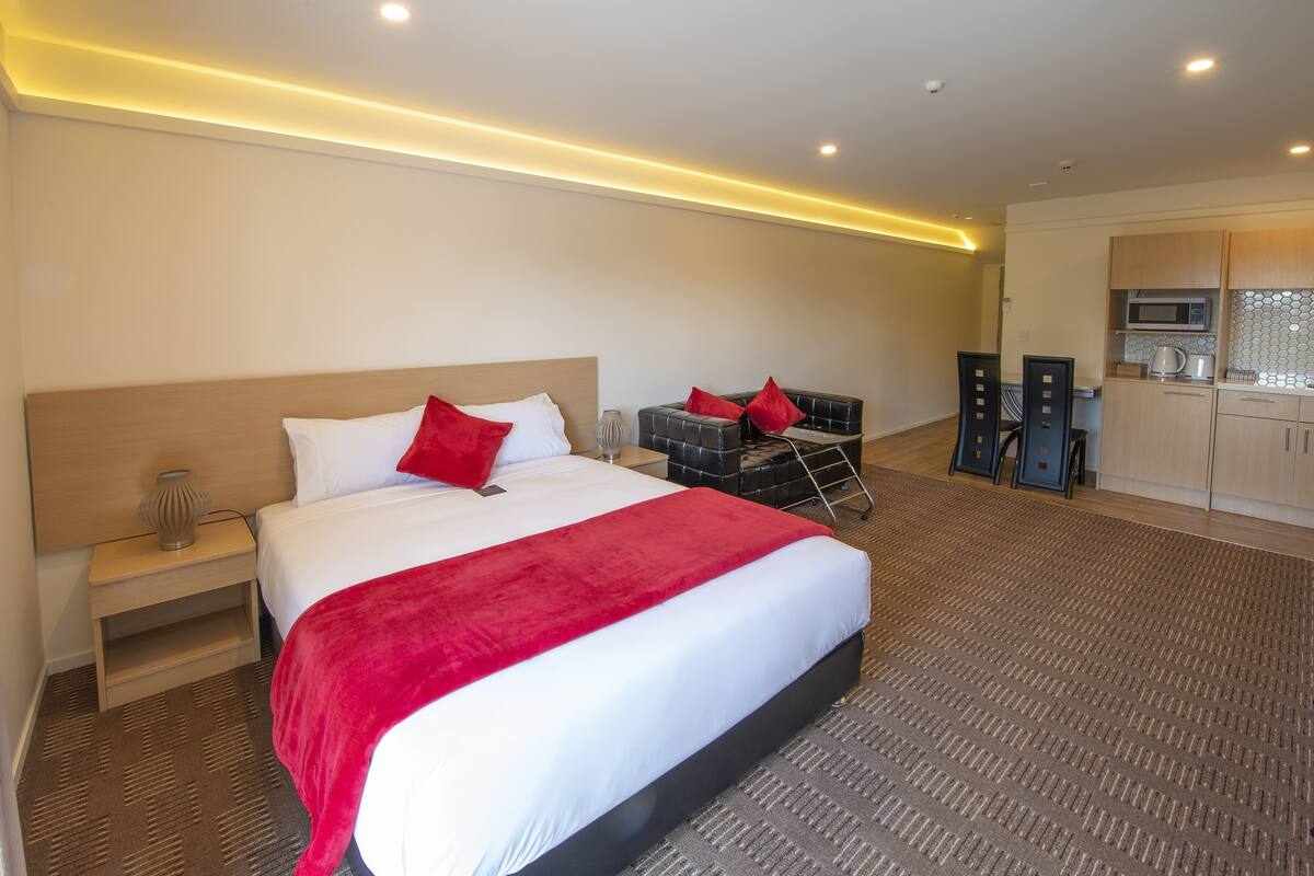 A hotel room with a large bed, a bedside table, a small table with a lamp, and a kitchenette area.
