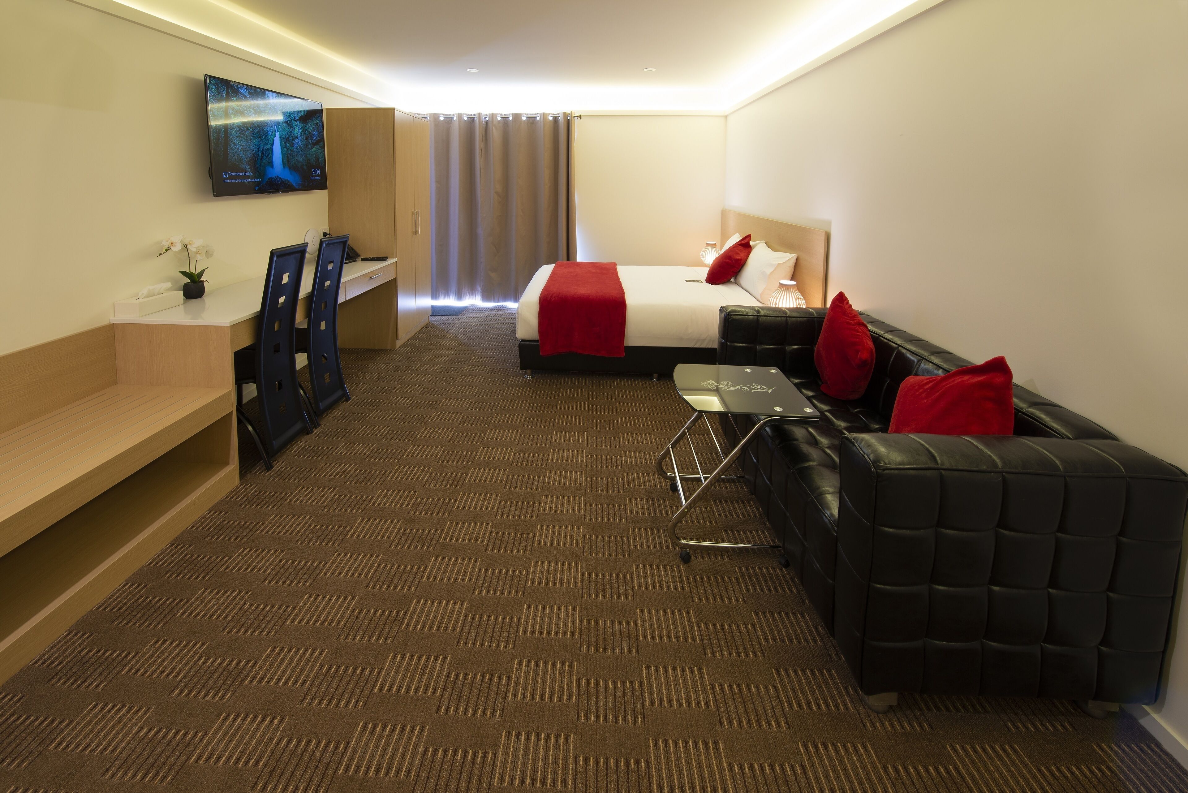 A hotel room with a bed, a TV mounted on the wall, a desk with chairs, and a sofa with red cushions.