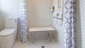 A bathroom with a hexagonal tile floor, a toilet, a shower with a handheld showerhead, and a bench.