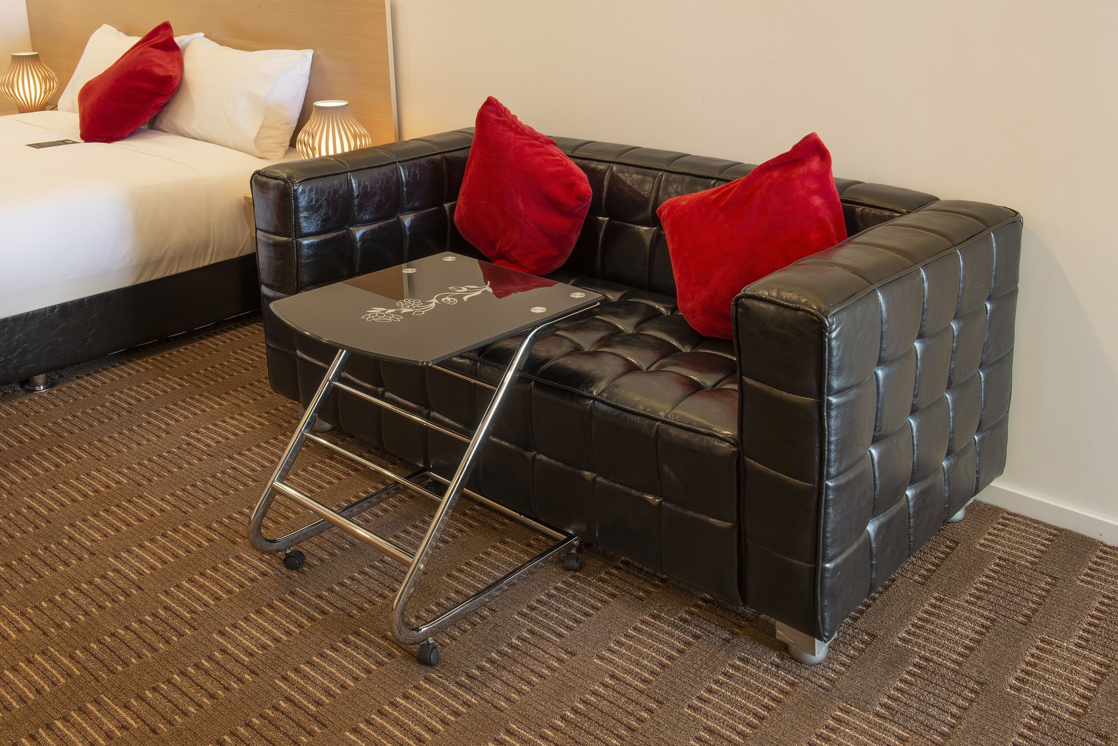 A hotel room with a black leather sofa, a small glass table, and a bed with white linens and red pillows.