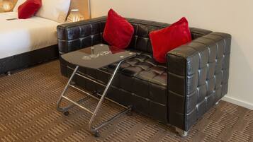 A hotel room with a black leather sofa, a small glass table, and a bed with white linens and red pillows.