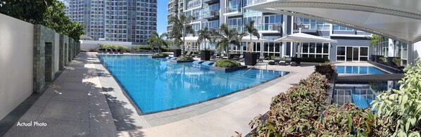 A modern hotel with a large rectangular pool, multiple lounge areas, and a view of high-rise buildings in the background.