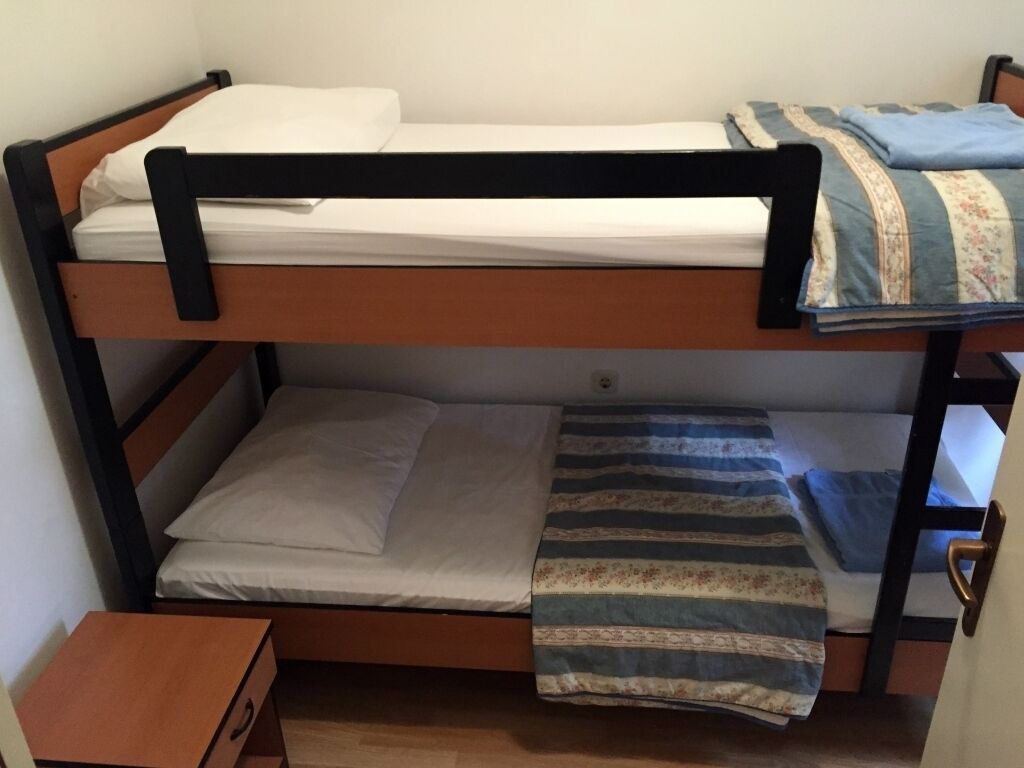 Un lit superposé à deux niveaux, chaque niveau étant équipé d’un matelas et d’oreillers. Une table de chevet en bois, dotée d’un tiroir, se trouve sous le lit du bas.