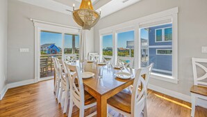 Zona pranzo con tavolo in legno e sedie bianche, ampie finestre con vista sugli edifici e un lampadario.