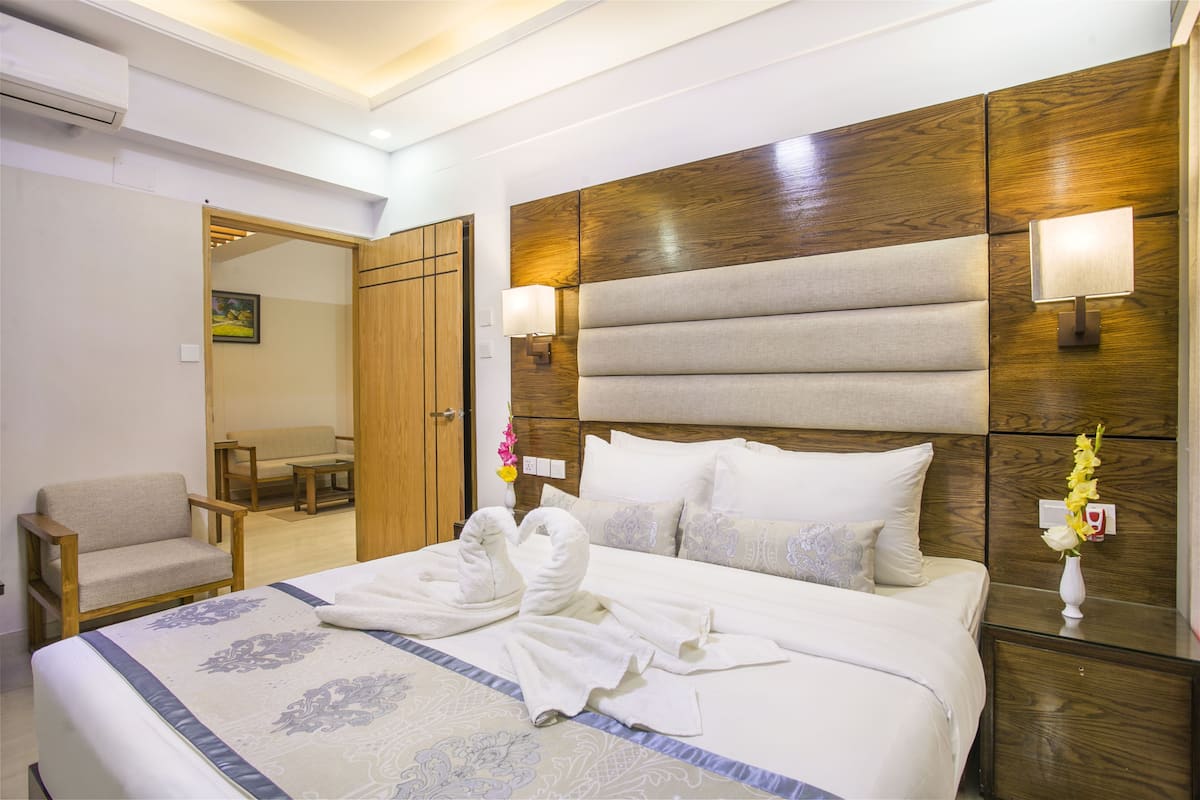A hotel room with a large bed, a wooden headboard, two bedside lamps, a nightstand with a vase of flowers, and a chair in the corner.