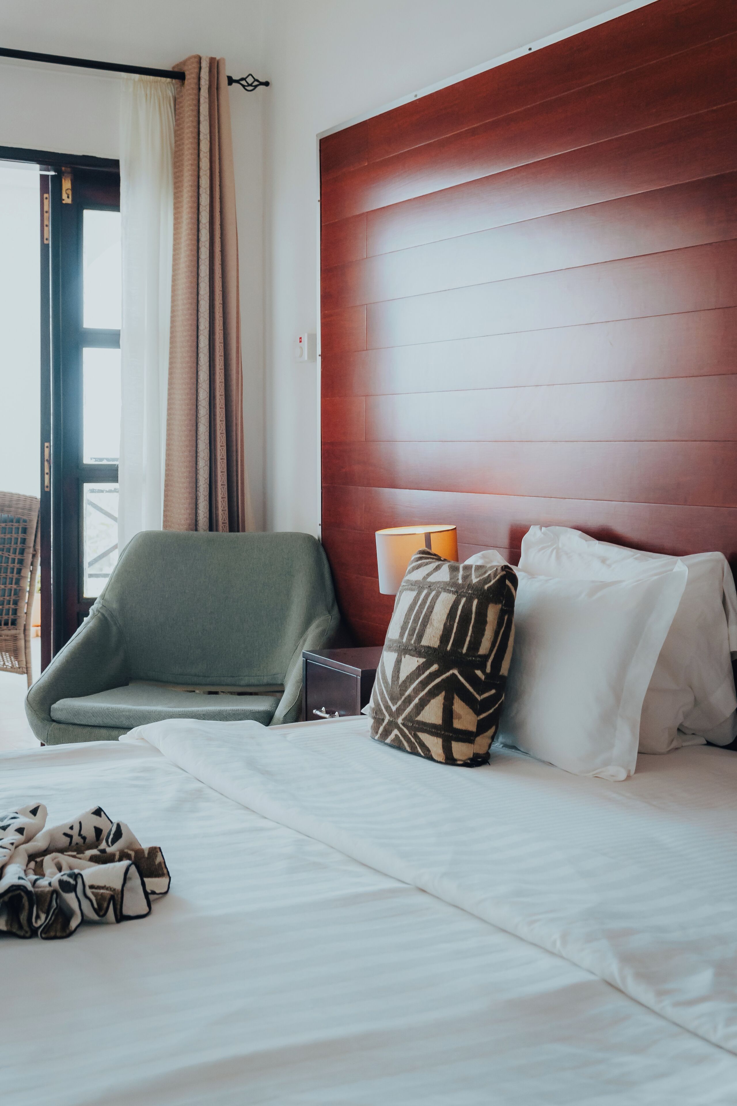 A hotel room with a bed, a chair, a nightstand, a lamp, and a view of the outdoors.