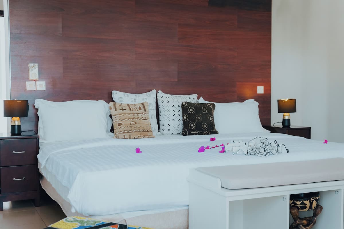 Een opgemaakt bed met gekleurde kussens, een nachtkastje met een lamp en een klein wit bankje.