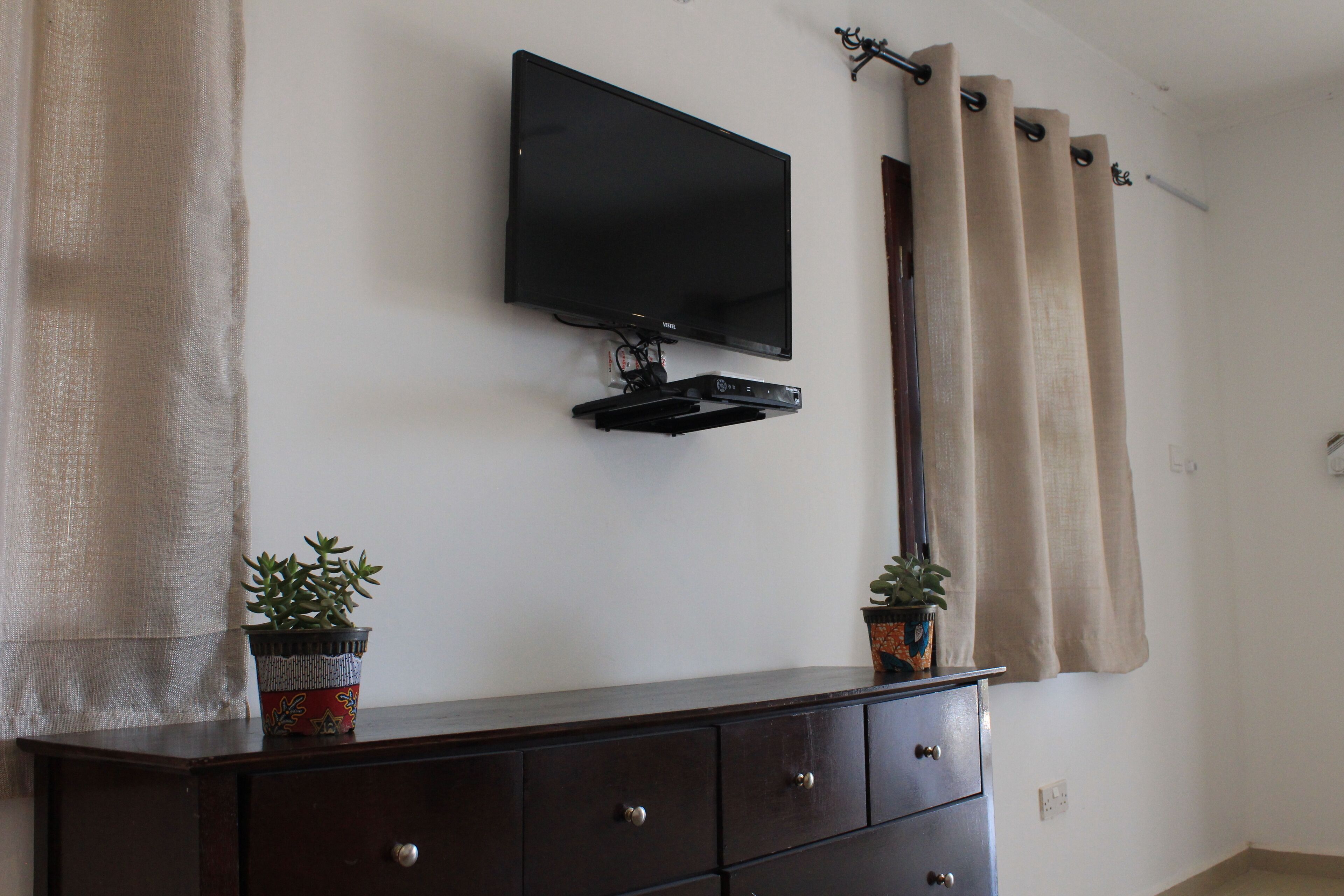 A room with a flat-screen TV mounted on the wall, a dark wooden dresser, and two potted plants.