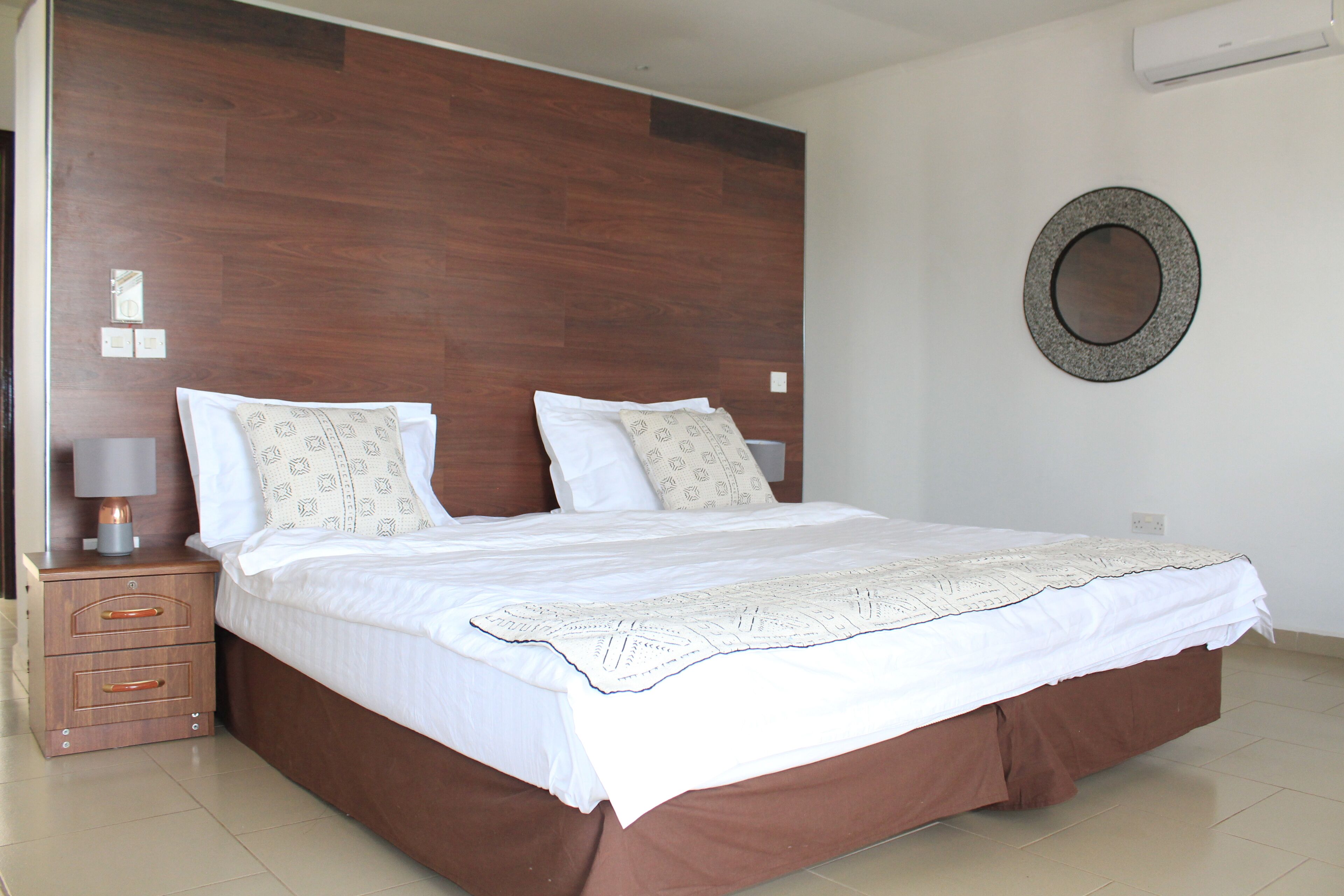A neatly made bed with a wooden headboard, a bedside table with a lamp, and a wall-mounted mirror.