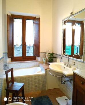 Un baño con bañera, lavamanos e inodoro. Hay una ventana con contraventanas de madera y una planta en maceta en el alféizar.