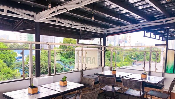An outdoor seating area with tables and wicker chairs, large windows with a view of greenery and buildings, and a ceiling with exposed beams and hanging light fixtures. - Chiasti Capsule Hostel - Adults Only (Cebu City)