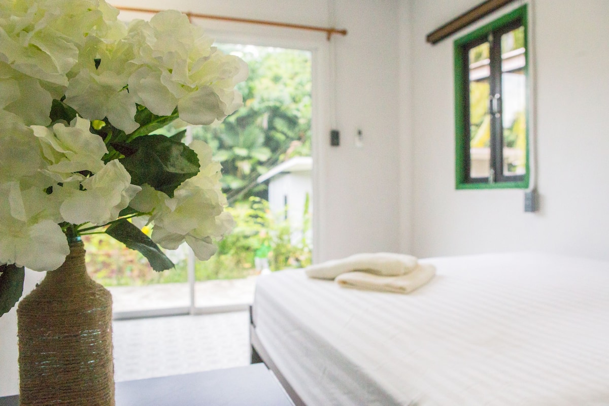 Quarto com cama, janela com moldura verde e um vaso com flores brancas.