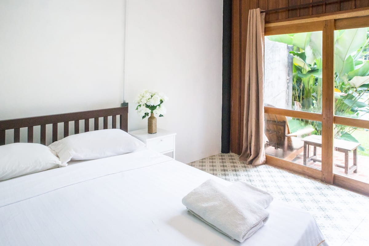 Quarto de hotel com uma cama, uma mesinha de cabeceira com um vaso de flores e vista para um jardim através de uma janela grande.
