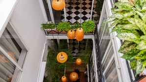 Un estrecho pasillo exterior con lámparas naranjas colgantes, plantas en macetas y un diseño de piso a cuadros.