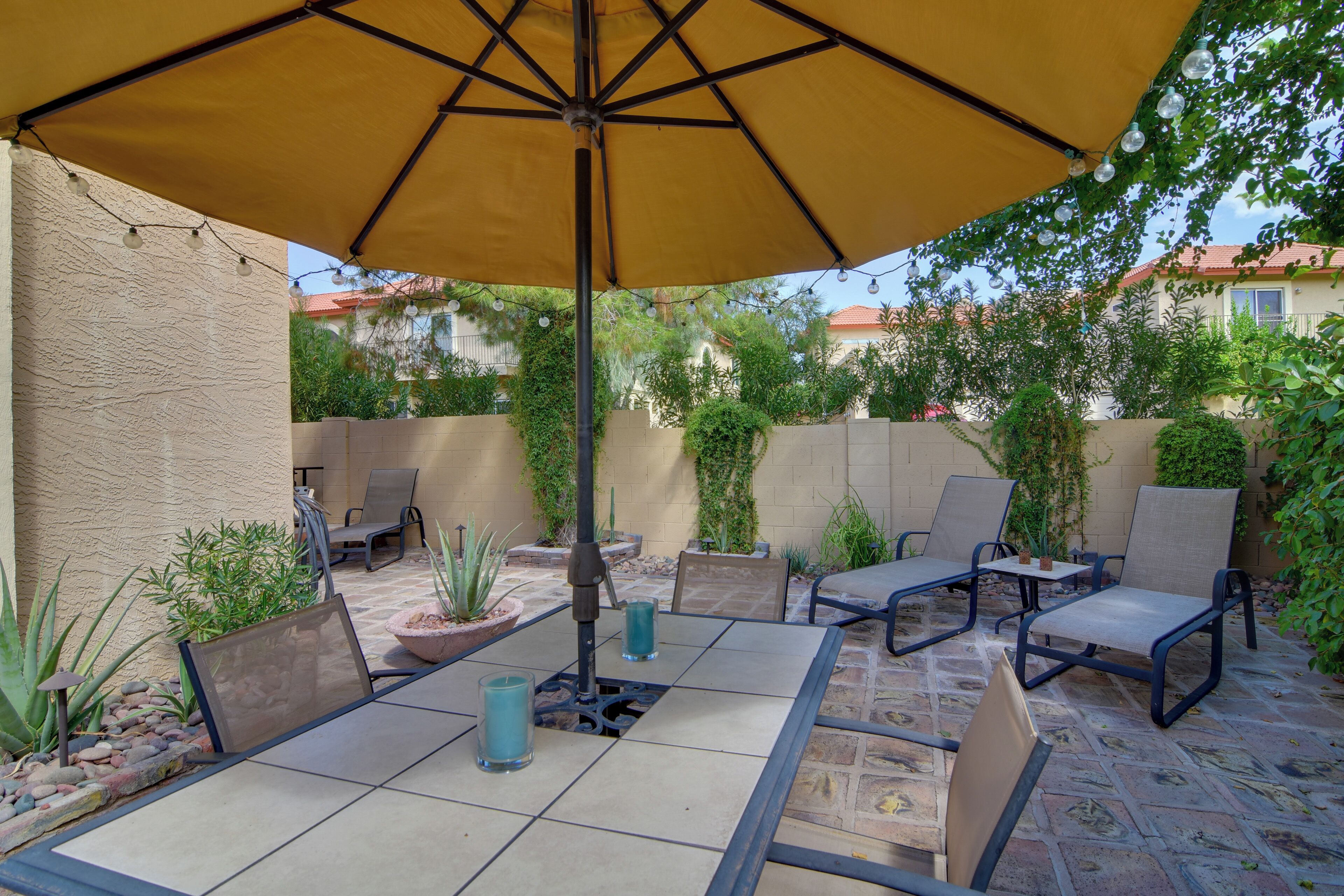An outdoor patio with a table and chairs under a large umbrella, surrounded by potted plants and a stone floor.