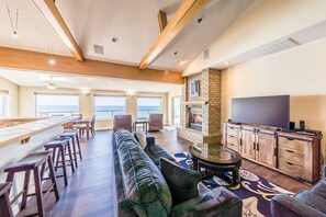 A spacious living area with a long dining table, comfortable seating, a fireplace, and a television. - Dramatic oceanfront views. Open high ceilings. Huge deck & walls of windows! (Lincoln City)