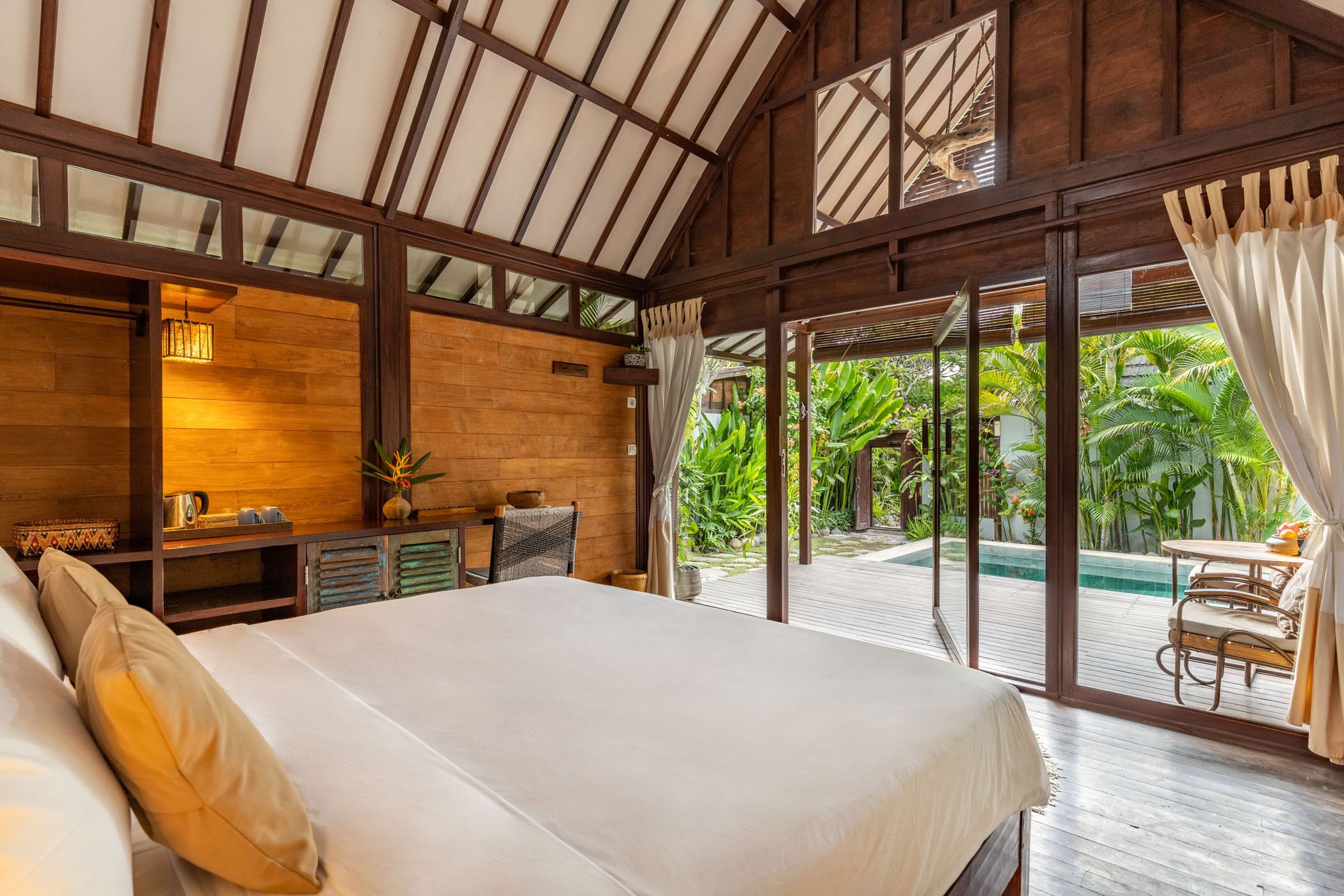 A spacious bedroom with a large bed, wooden walls, a desk with a lamp, and a view of a pool and tropical garden.