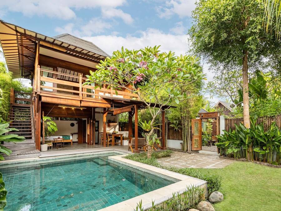 A two-story wooden house with a swimming pool, covered outdoor seating, and lush greenery.