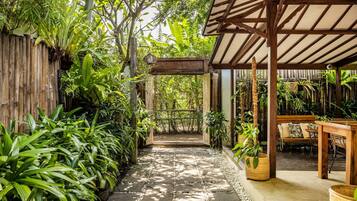 A covered outdoor area with a stone pathway, wooden furniture, and lush greenery.
