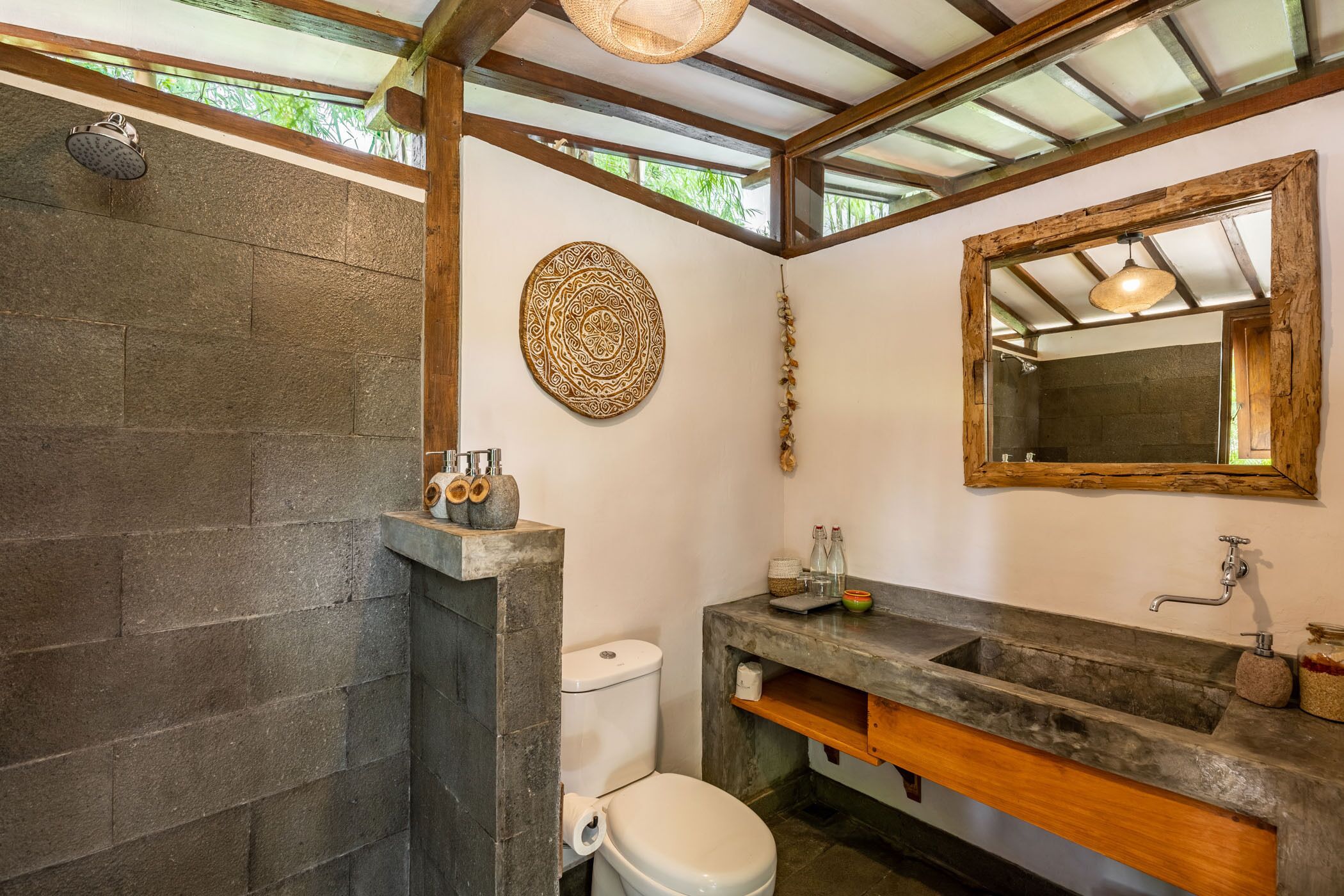 A bathroom with a stone shower, a toilet, a sink with a wooden vanity, and a decorative wall hanging.