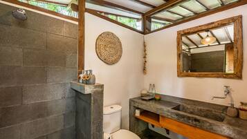 A bathroom with a stone shower, a toilet, a sink with a wooden vanity, and a decorative wall hanging.