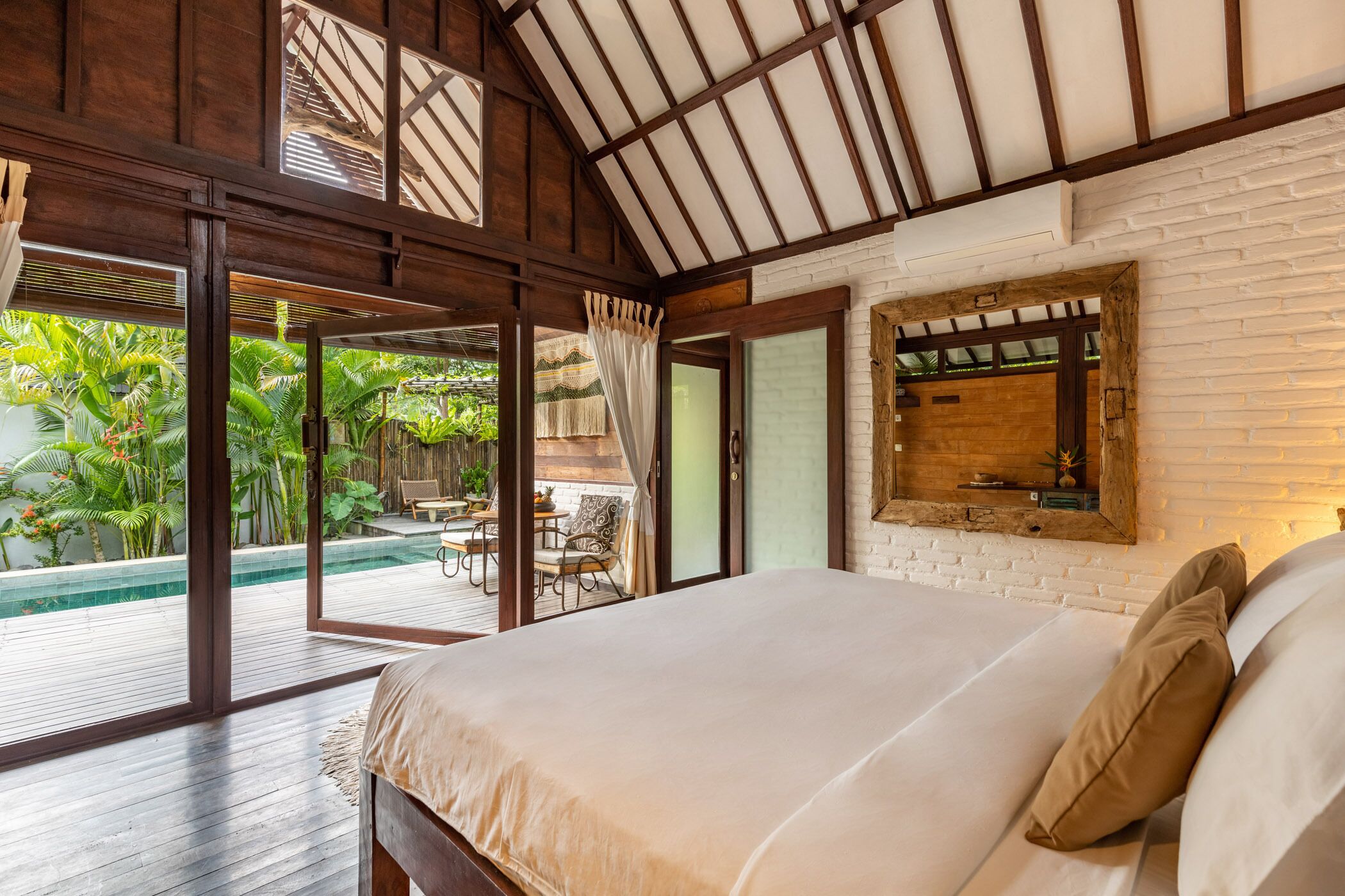 Ein Schlafzimmer mit einem großen Bett, Holzbalkendecke, einer Ziegelwand und Blick auf einen Pool und Grünflächen.